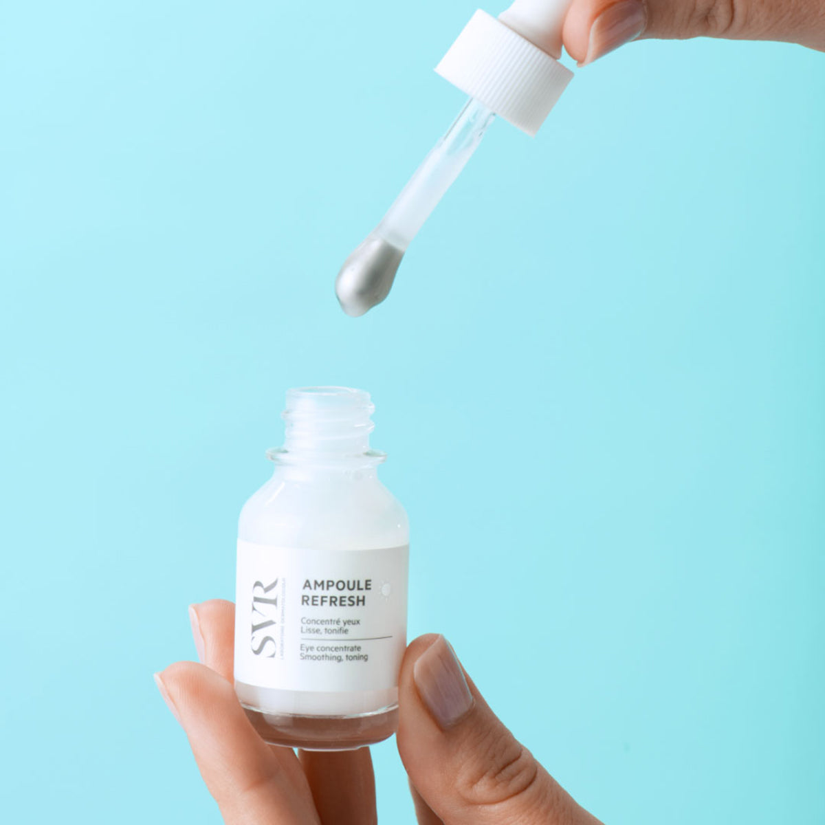 Serum pod oczy SVR Ampoule Refresh w szklanej buteleczce z metalowym aplikatorem w dłoni na jasnoniebieskim tle.