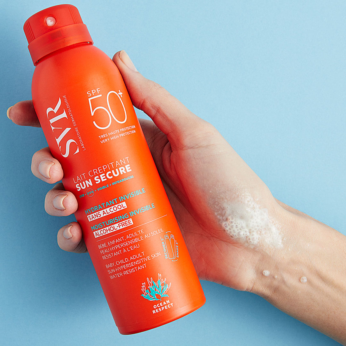 Dłoń trzymająca pomarańczowy spray SVR Sun Secure SPF50+ z pianką na skórze na niebieskim tle.