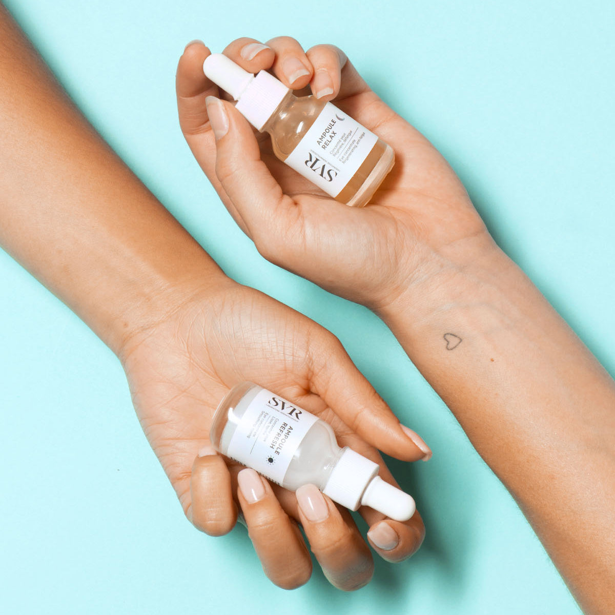 Dwie dłonie trzymające szklane buteleczki serum SVR Ampoule Refresh i Relax z białymi zakrętkami na jasnoniebieskim tle.