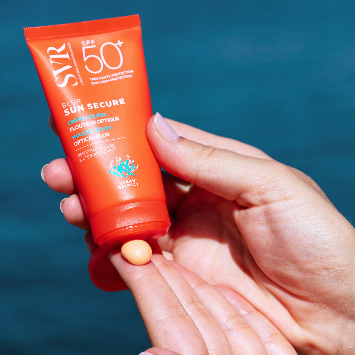 Pomarańczowa tubka kremu SVR Sun Secure Blur SPF50+ z brzoskwiniową konsystencją musu wyciskaną na dłoń.