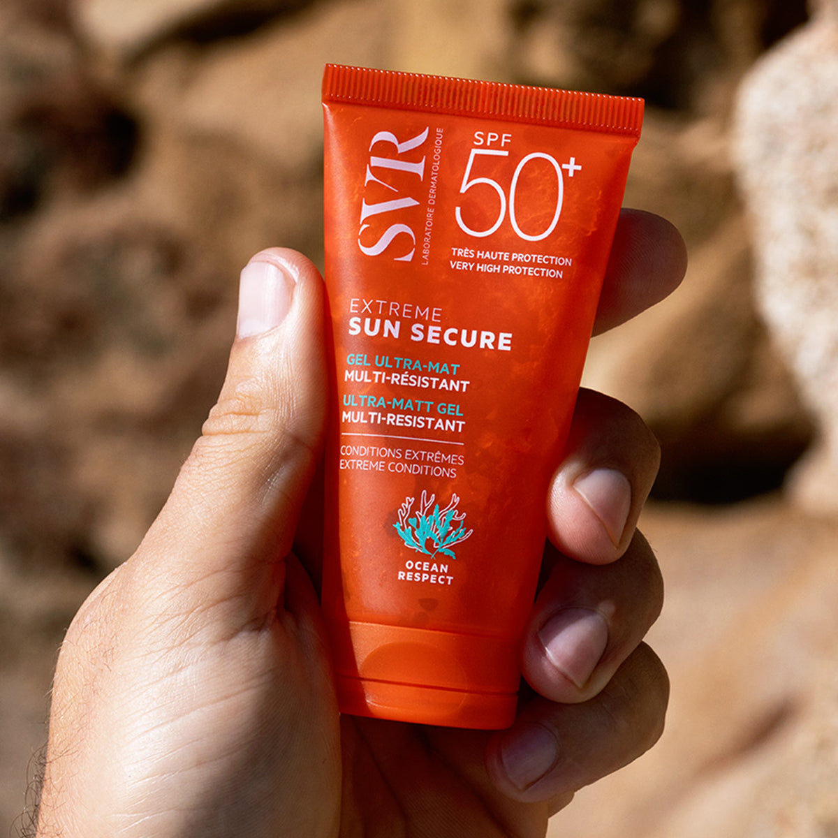 Pomarańczowa tubka żelu ochronnego SVR SUN SECURE Extreme SPF50+ trzymana w dłoni na tle skał.