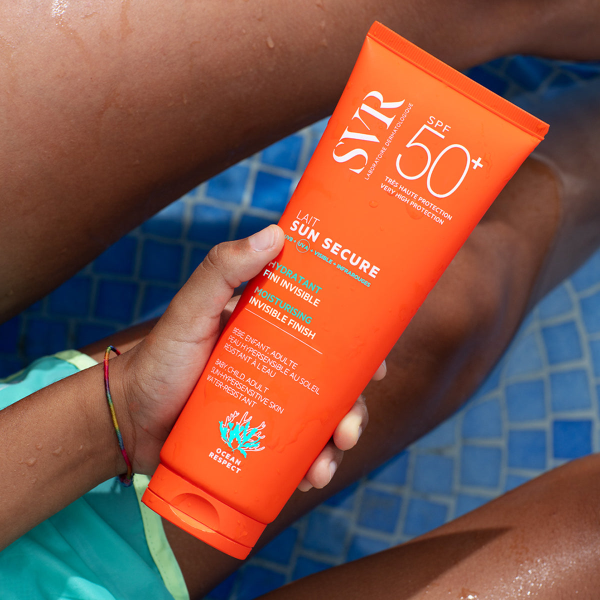 Pomarańczowa tubka mleczka przeciwsłonecznego SVR Sun Secure SPF50+ trzymana w dłoni na tle basenu.