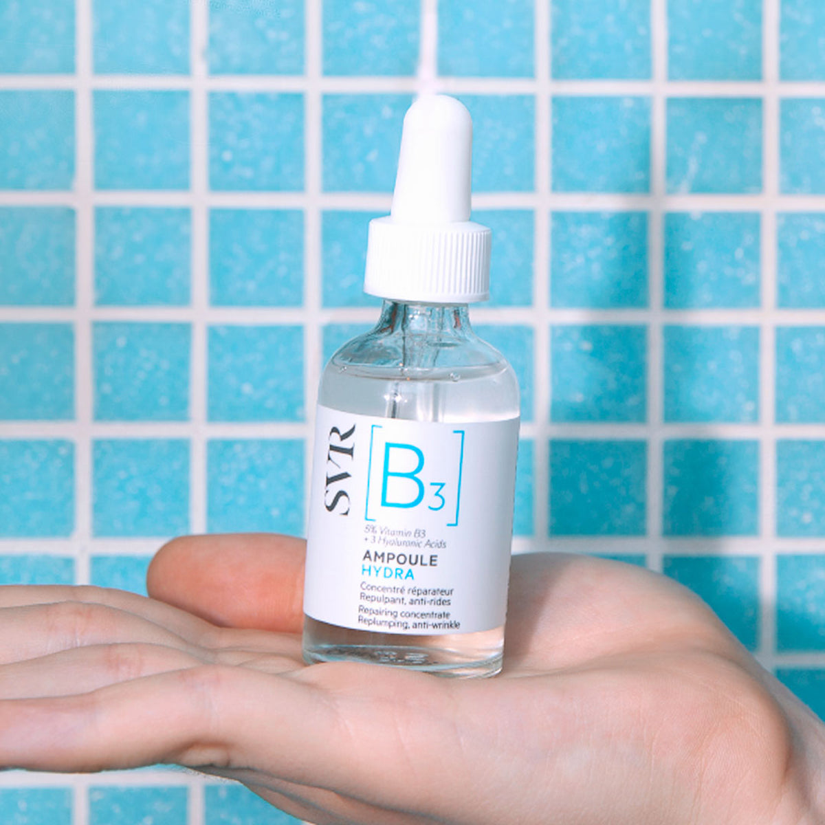 Szklana buteleczka serum SVR Ampoule Hydra z pipetą trzymana w dłoni na tle niebieskich kafelków.