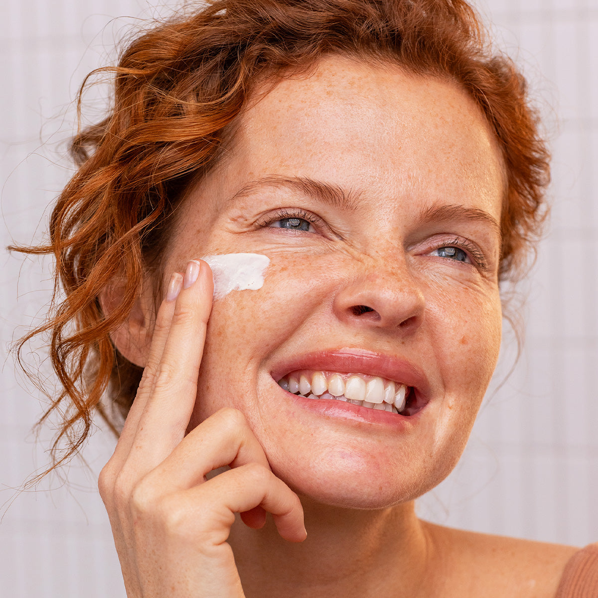Smiling woman with red hair applying white SUN SECURE SPF50+ fluid sunscreen to her cheek against a tiled background.