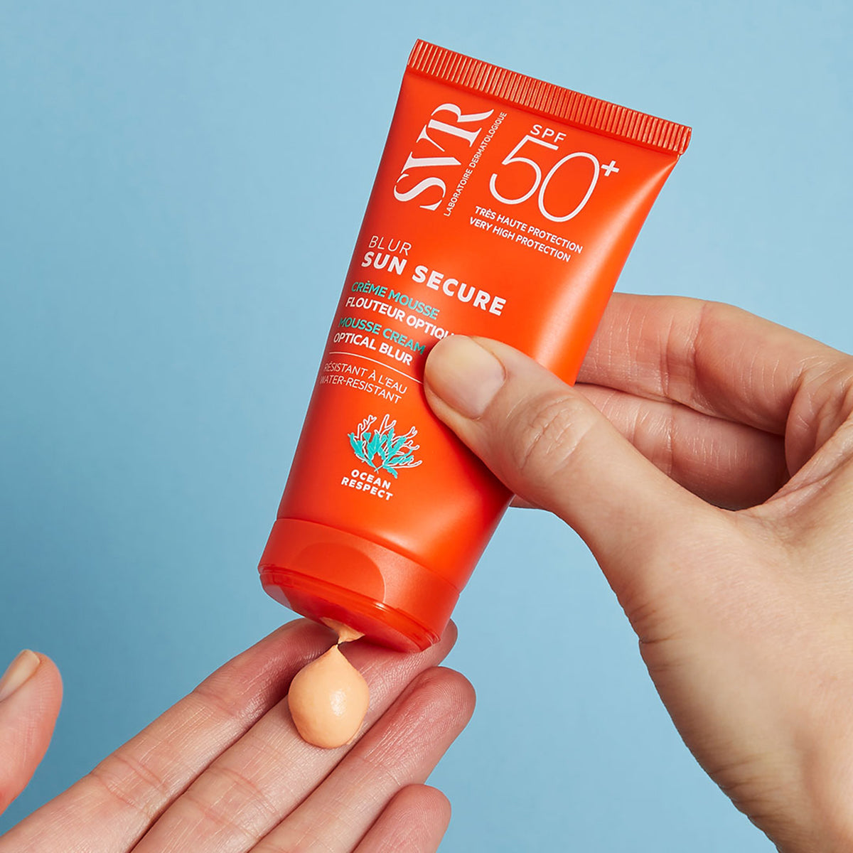 Pomarańczowa tubka kremu SVR Sun Secure Blur SPF50+ z brzoskwiniową teksturą wyciskaną na dłoń na niebieskim tle.