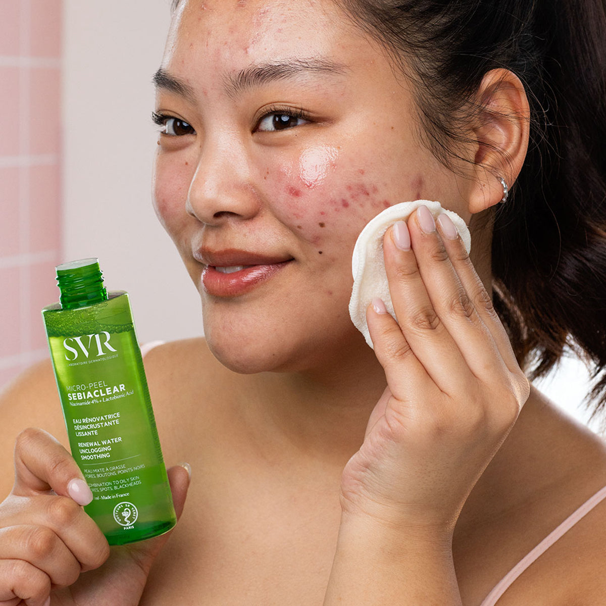 Kobieta nakłada wacikiem esencję SVR Sebiaclear Micro-Peel z zielonej, przezroczystej butelki na skórę z niedoskonałościami.