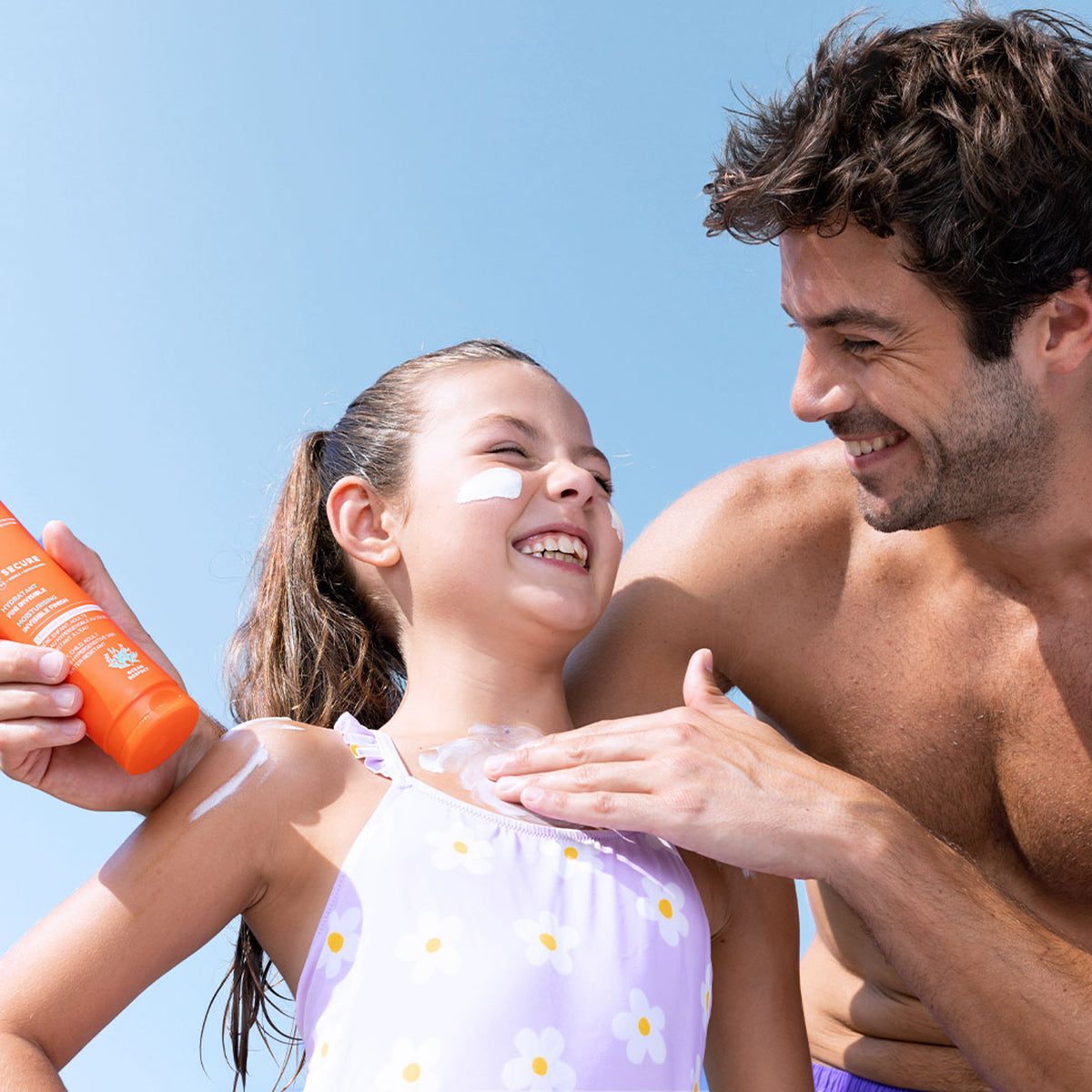 Ojciec nakłada pomarańczową tubkę mleczka przeciwsłonecznego SVR Sun Secure SPF50+ na skórę uśmiechniętej dziewczynki.