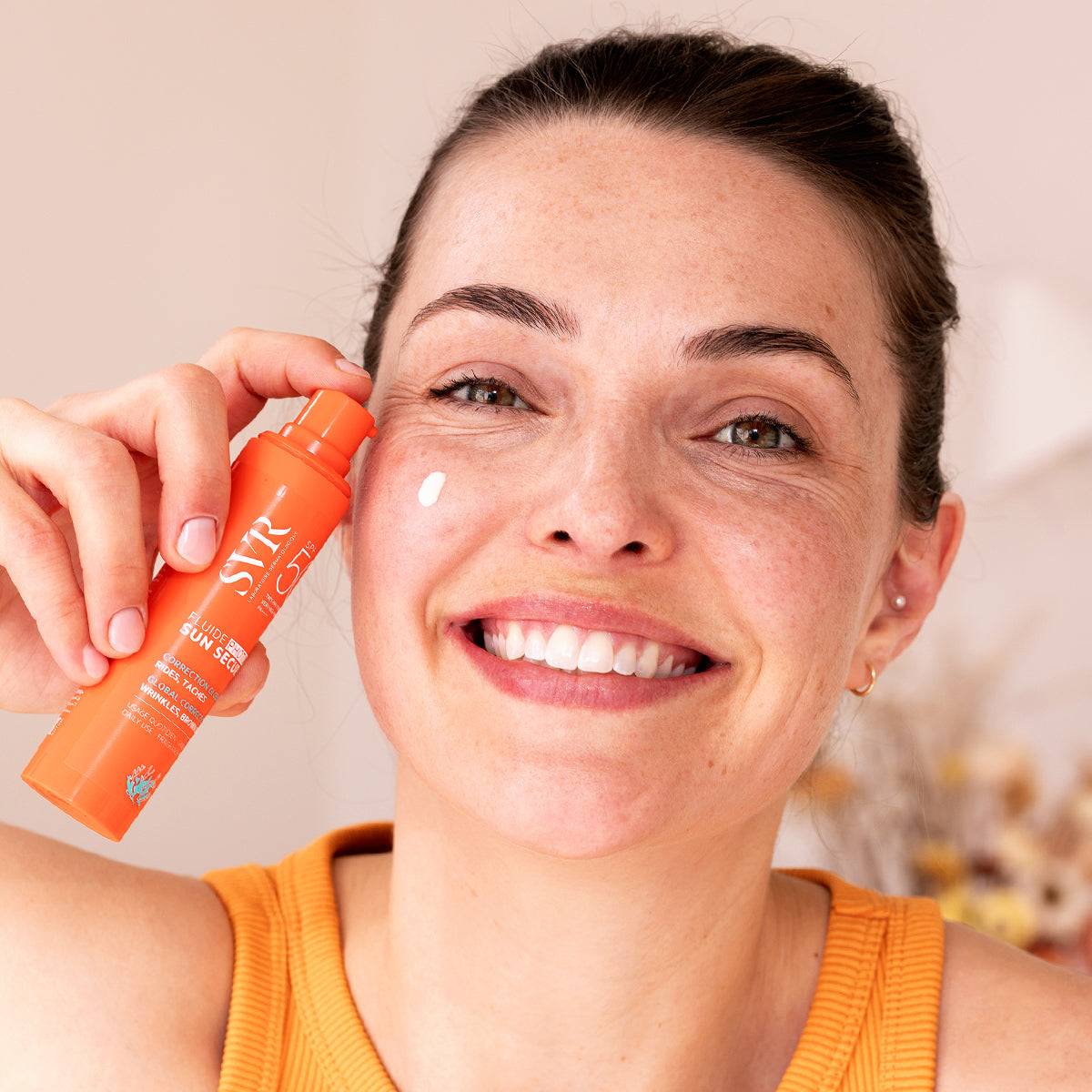 Smiling woman holding an orange SVR SUN SECURE SPF50+ pump bottle with a dab of cream on her cheek.