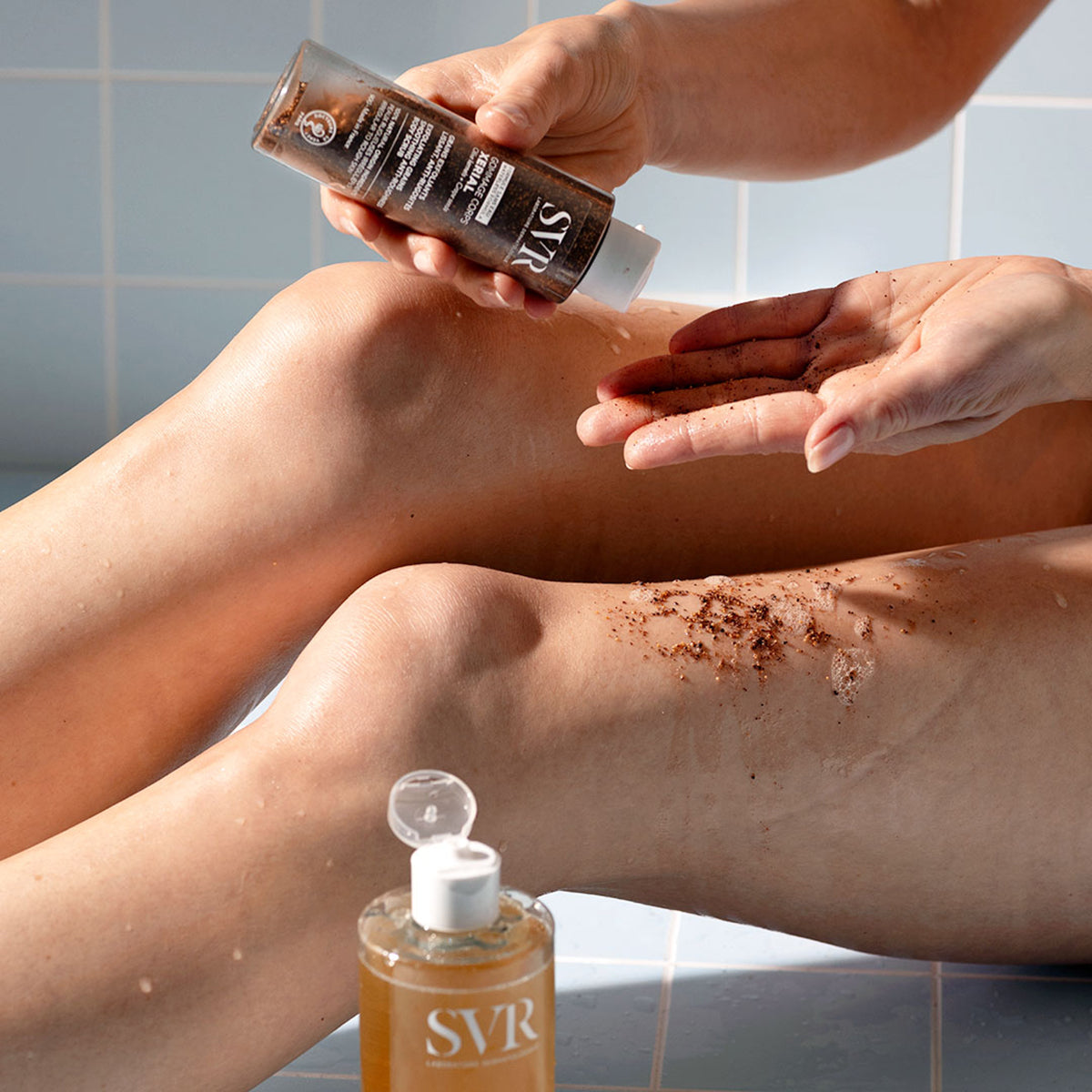 Osoba nakłada peeling SVR XERIAL Gommage z drobinkami pestek oliwek i winogron na nogę w łazience.