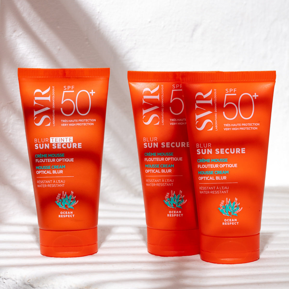 Trzy pomarańczowe tubki kremu SVR SUN SECURE Blur SPF50+ z napisem Ocean Respect na jasnym, cieniowanym tle.