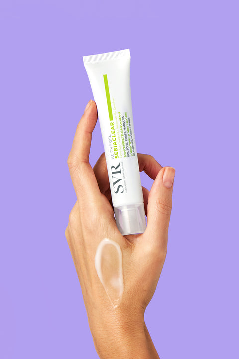 Hand holding a white tube of SVR Sebiaclear Active Gel with a translucent gel texture swatch on the skin, purple background.