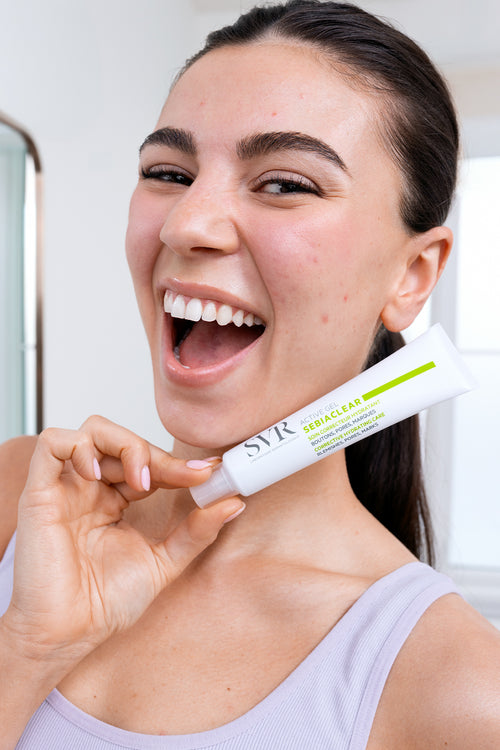 Une femme souriante tenant un tube de gel correcteur SVR Sebiaclear Active pour le soin des peaux à imperfections.