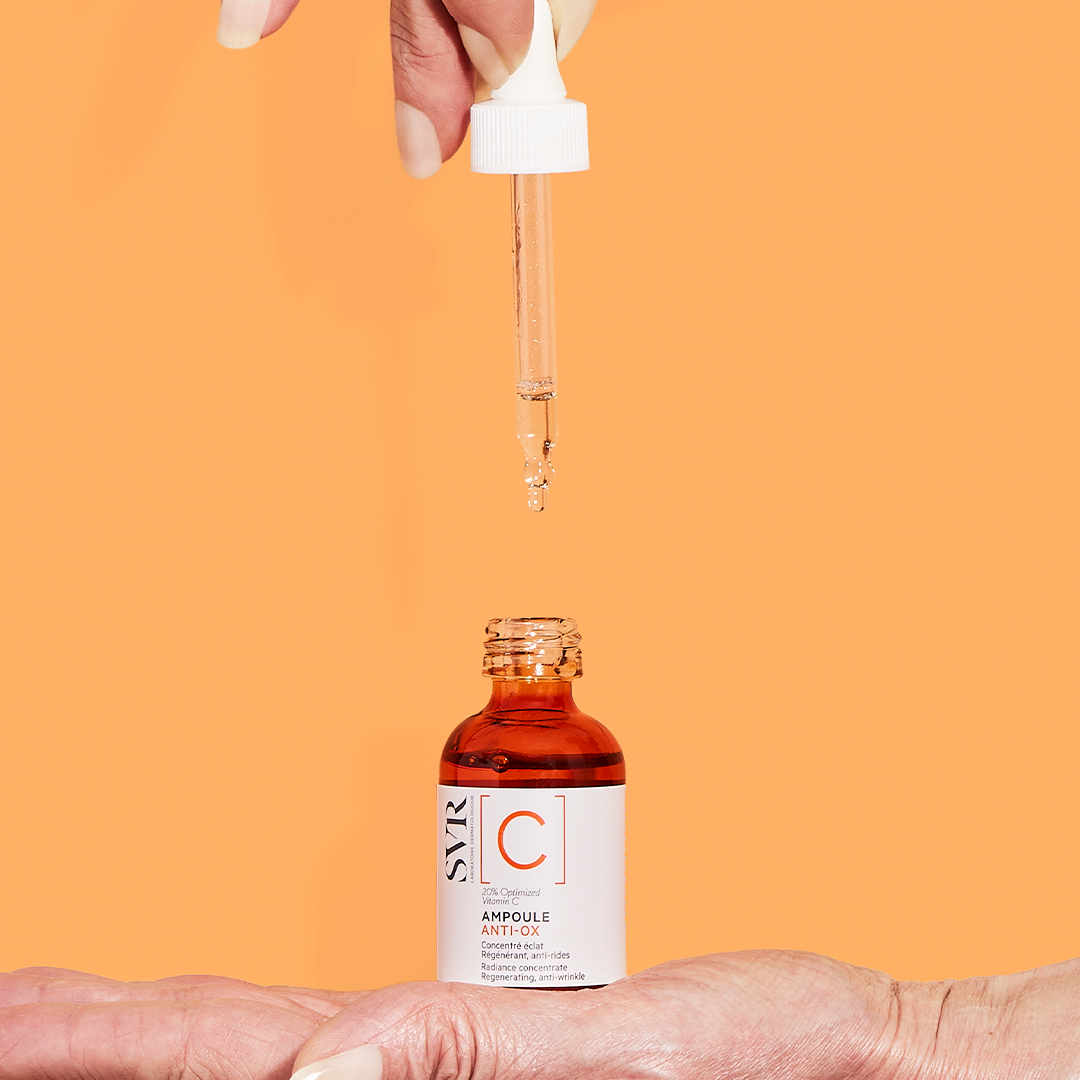 SVR Ampoule [C] anti-oxidant serum in an amber glass bottle with a clear dropper held over a hand on an orange background.