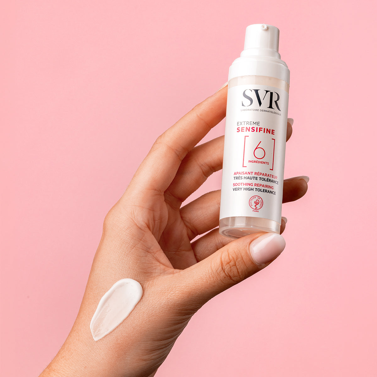 Hand holding a white SVR Sensifine Extreme pump bottle with a dollop of white cream on the skin against a pink background.